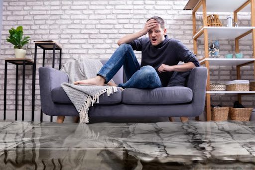 A male homeowner sits on his couch in the living room which is flooding with water from his plumbing. He looks stressed and needs an expert plumber in Marshfield, MO.