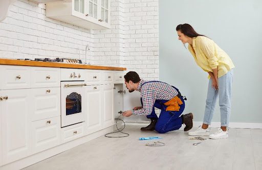 A professional male plumber crouches on the floor in a modern white kitchen while using a drain cable to clean a clogged sink pipe as a female homeowner watches in Strafford, MO.