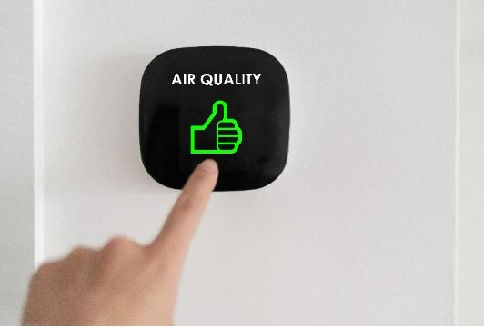 Hand pointing at a circular air quality monitor displaying a green thumbs-up icon on a white wall.
