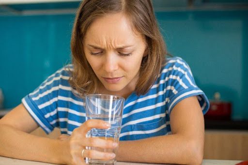 A woman in a blue and white shirt looking at her glass of water with a confused look due to the off smell of the water from her kitchen faucet in Springfield, MO.