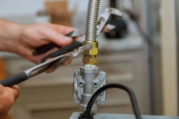 Person using pliers to tighten a flexible gas line connection on a metal valve assembly in a kitchen-like setting.