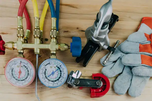 HVAC toolkit on a wooden surface with pressure gauges, colored hoses, a pipe cutter, wrench, gloves, and a silver tool.