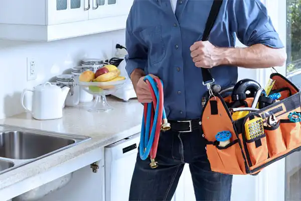 A person holding a tool bag and colorful hoses stands in a kitchen near a countertop with fruit and jars.