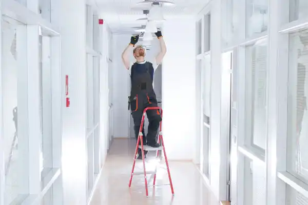 Person standing on a ladder, wearing overalls and a cap, adjusting ceiling lights in a hallway with glass-panelled doors.