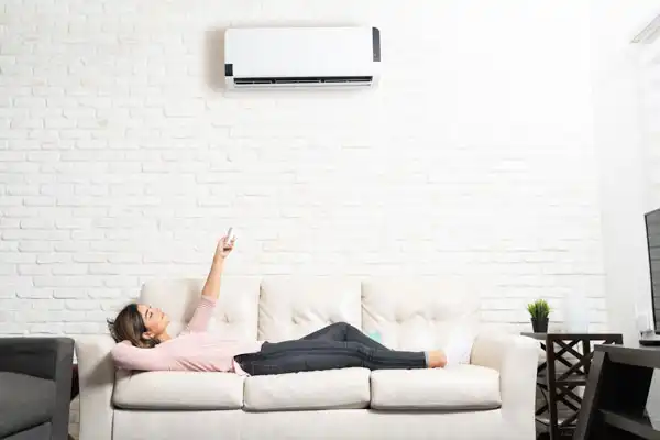 A woman relaxes on a sofa, using a remote to operate an air conditioner mounted on a white brick wall.