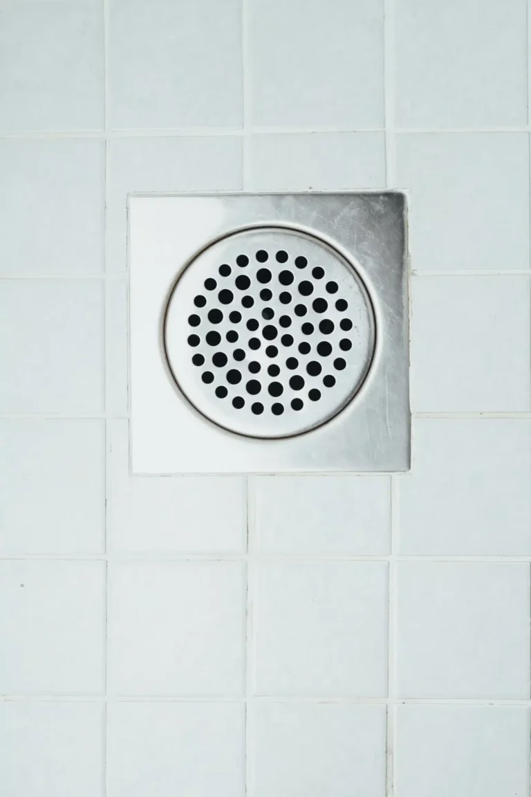A square shower drain embedded in white tiled flooring, with a circular metal grate featuring multiple holes.