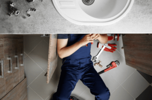 Person lying on the floor under a sink fixing plumbing. Tools and parts are scattered around.