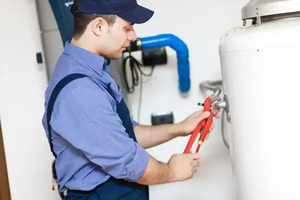 A plumber in a blue uniform and cap adjusts a fitting on a white water heater using red-handled pliers.