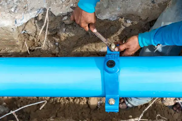 Person using a wrench to tighten a bolt on a blue underground pipe in a dirt trench.