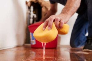 A person kneels on a wooden floor, using a yellow sponge to soak up water, with a red bucket in the background.