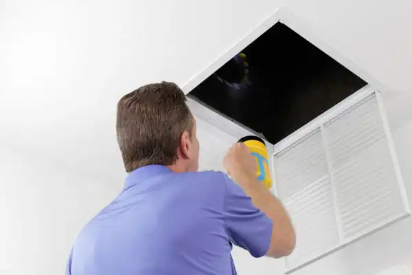 Person in blue shirt shining a flashlight into an open ceiling vent.