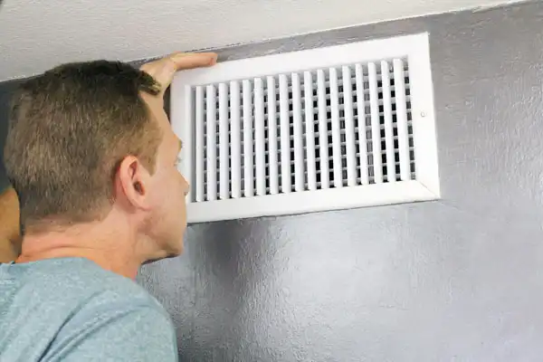 A person inspecting a white air vent on a gray wall.