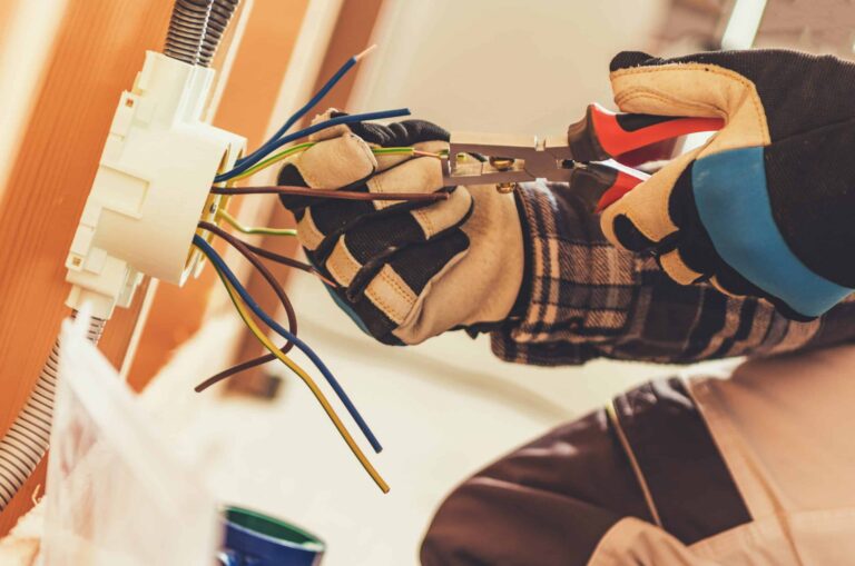 A person wearing gloves is using pliers to work on electrical wires protruding from a wall socket.