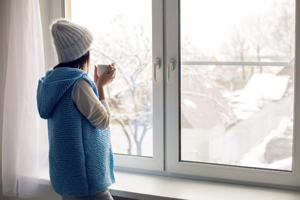Person wearing a blue knit vest and gray beanie stands at a window holding a mug, looking out at a snowy landscape with trees and rooftops.
