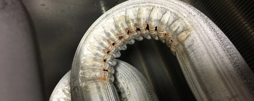 Close-up of a corroded, bent metallic pipe with visible rust spots and mineral deposits, likely part of an HVAC or refrigeration system.
